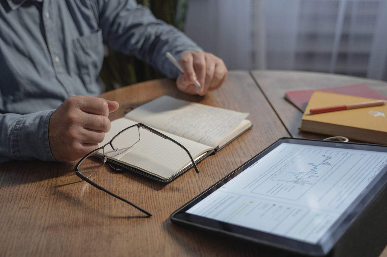 A man writes in a notebook beside a tablet on a wooden table.